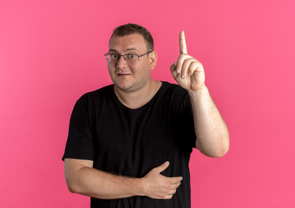 看戴眼镜的超重男子身穿黑色t恤 微笑着展示食指 站在粉红色的墙上有一个新的好主意伟大穿显示
