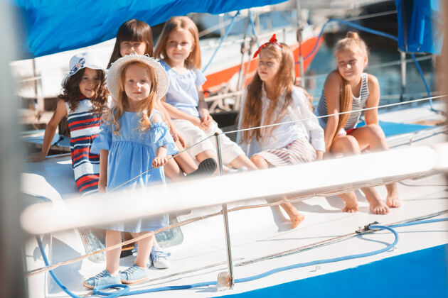 船孩子们在游艇上喝着橙汁青少年或少女们在蓝天下户外五颜六色的衣服孩子们的时尚 阳光明媚的夏天 河流和假日的概念女孩水手船