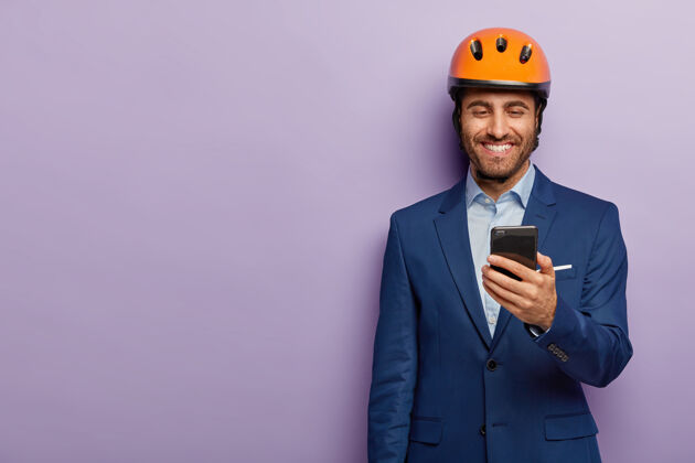 男人开朗的建筑师戴着橙色安全帽 正装 在工作间隙有空余时间 在智能手机上收到信息 很高兴收到薪水凝视建筑高兴