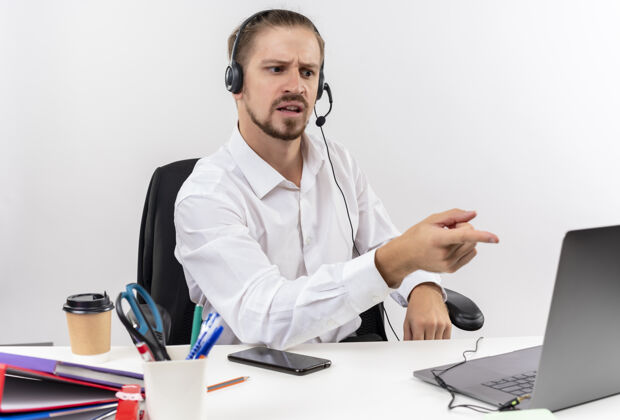 坐着穿着白衬衫 戴着耳机 拿着麦克风的帅哥坐在白色背景下的办公桌旁 看着困惑的笔记本电脑困惑工作白色