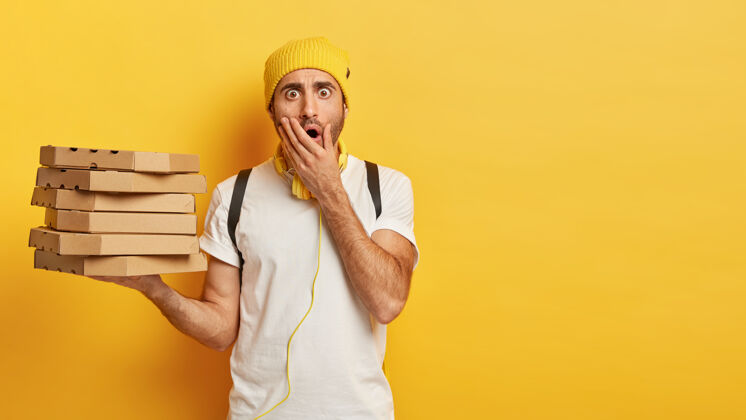 堆年轻震惊的男送货员的画像拿着一叠披萨盒 穿着随意 捂着张开的嘴 靠着黄色的墙站着年轻人男性纸板