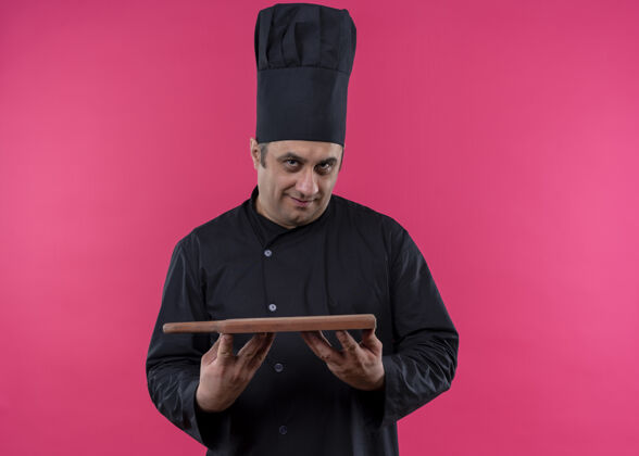 剪男厨师身穿黑色制服 头戴厨师帽 手持切割木板 自信地站在粉色背景上木板制服自信