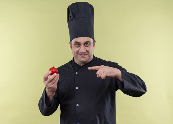 烹饪男厨师身穿黑色制服 头戴厨师帽 手指指着番茄微笑着看着站在绿色背景上的摄像机微笑看站