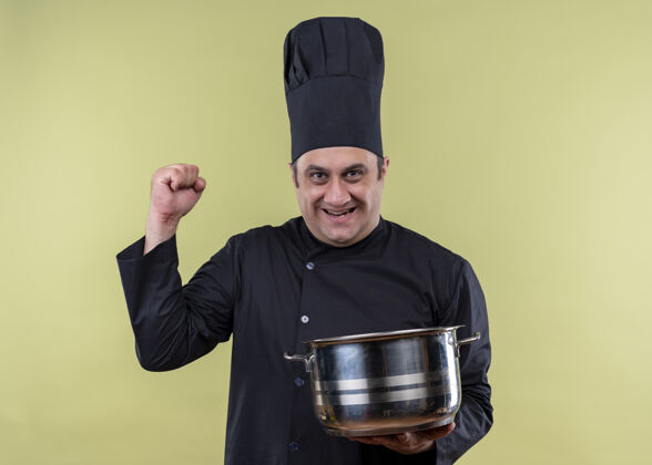 烹饪男厨师身穿黑色制服 头戴厨师帽 手持平底锅 微笑着看着镜头 兴奋地握紧拳头站在绿色背景上拳头看着厨师