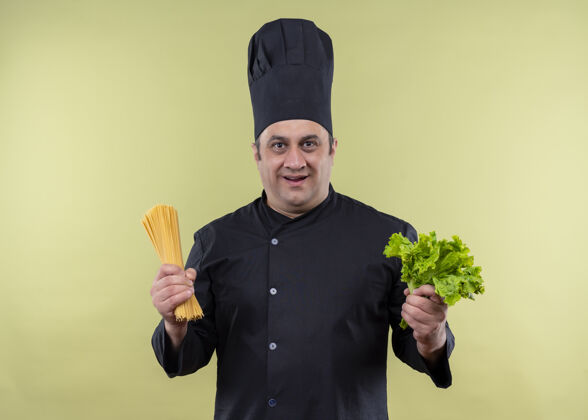 制服男厨师身穿黑色制服 头戴厨师帽 手里拿着生意大利面和新鲜莴苣 站在绿色背景上 看上去既高兴又惊讶帽子看烹饪