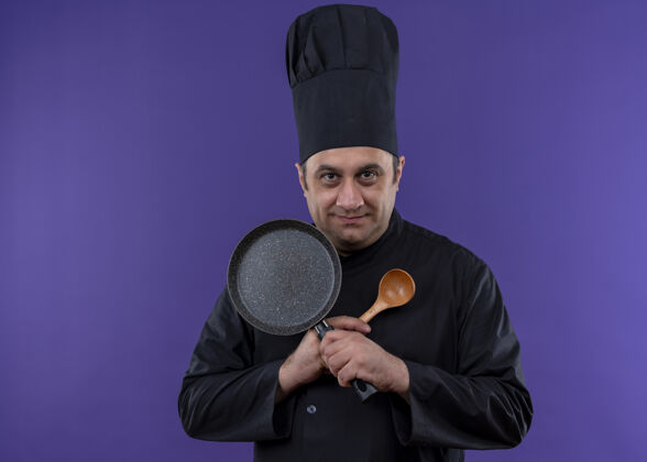 薯条男厨师身穿黑色制服 头戴厨师帽 手拿煎锅和木勺 双手交叉看着镜头 站在紫色背景下微笑着男拿着平底锅