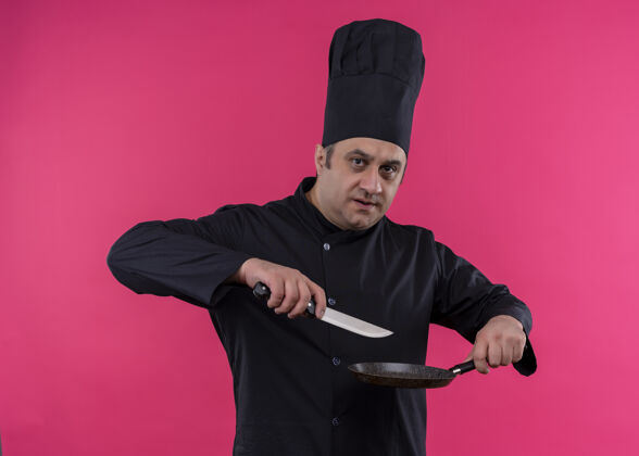 帽子男厨师身穿黑色制服 头戴厨师帽 拿着平底锅和刀 严肃的脸站在粉色背景下看着镜头烹饪拿着平底锅
