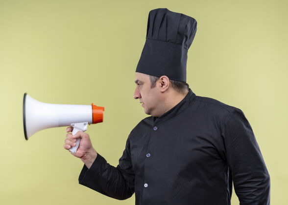 靠边男厨师身穿黑色制服 头戴厨师帽 表情严肃 手持扩音器站在绿色背景上看拿着厨师