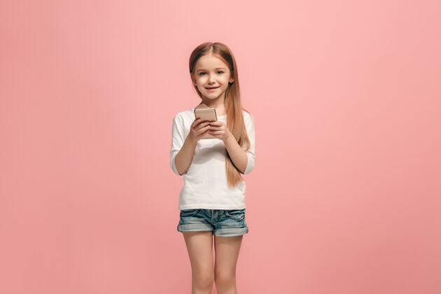 教育快乐的少女站着 微笑着用手机在时尚的粉色工作室背景上美丽的女性半身像人类的情感 面部表情的概念青少年手机欢呼