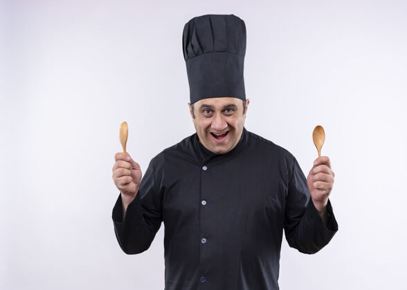白男厨师身穿黑色制服 头戴厨师帽 手持两个木勺 站在白色背景下 面带微笑 看着镜头男烹饪木头