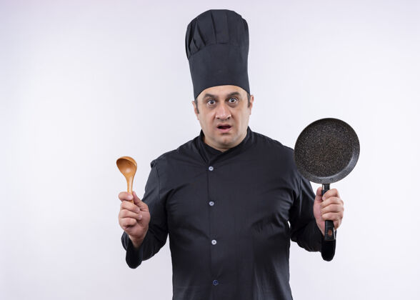 白男厨师身穿黑色制服 头戴厨师帽 拿着平底锅和木勺 站在白色背景下惊讶地看着镜头惊喜厨师勺子
