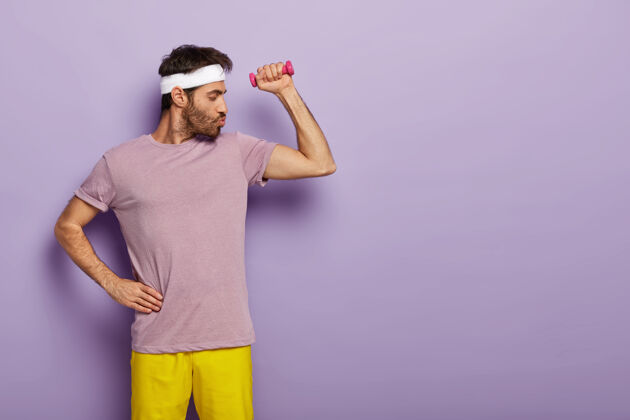 紫色强壮有力的男人 浓密的鬃毛 穿着随意 一只手放在腰上 用哑铃做运动 享受规律的训练室内力量教练