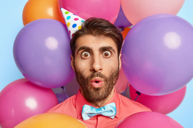 气球令人印象深刻的生日男人的特写镜头盯着的眼睛 有难以置信的面部表情 戴着纸锥帽 优雅的粉红色衬衫和领结尴尬惊奇站