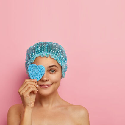 外观高兴的女模特用心形海绵捂着眼睛 戴着淋浴帽 赤裸着站在粉红色的墙上 擦着化妆品 有卫生程序 关心脸部皮肤 室内模特欧式化妆品人