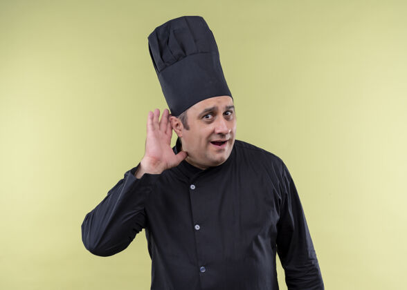 男性男厨师身穿黑色制服 手拿厨师帽 站在绿色背景下 试图倾听别人的对话尝试帽子有人