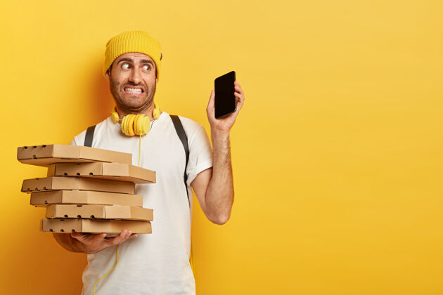 披萨照片中困惑的送货员端着比萨饼盒 拿着现代化的智能手机 一次接到很多电话和订单 穿着休闲装 靠着黄色的墙站着盒子男性愤怒