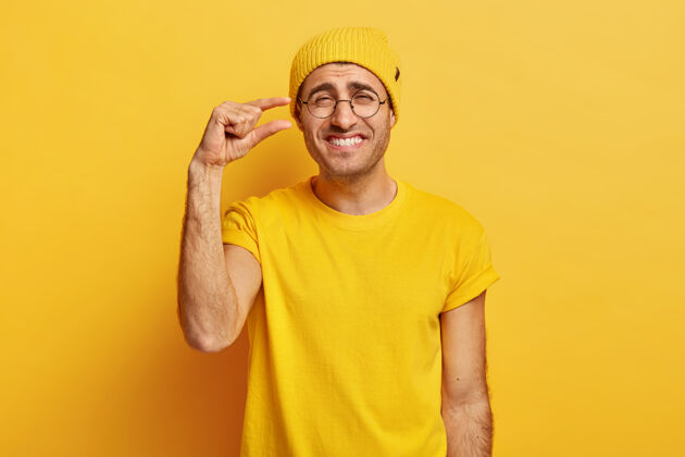 鬃毛快乐的男人穿着休闲的黄色衣服 做着微小的手势 露出很少的东西 高兴地微笑 表情愉快 戴着透明的眼镜胡茬眼镜微笑