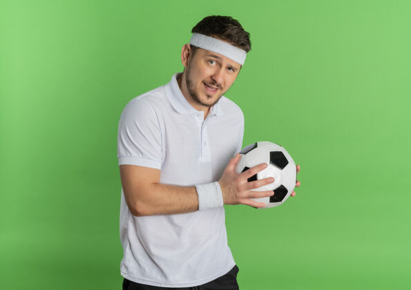 抱着身穿白衬衫 头戴足球带的年轻健身男子站在绿色背景下 兴高采烈地微笑着看着摄像机衬衫男士看