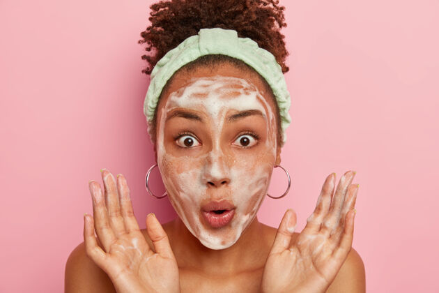 清洁惊呆了的女人在淋浴后洗脸 脸上有肥皂泡 抬起手掌 眼睛盯着器 戴着头带 模特在粉红色的墙上生活方式的形象肥皂赤膊上阵浴室
