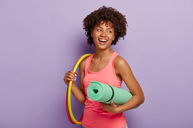姿势运动型苗条女士 健康的深色皮肤 非洲发型 呼啦圈练习 提着卷起的垫子 穿着粉色背心 有着牙齿般的微笑微笑快乐女性