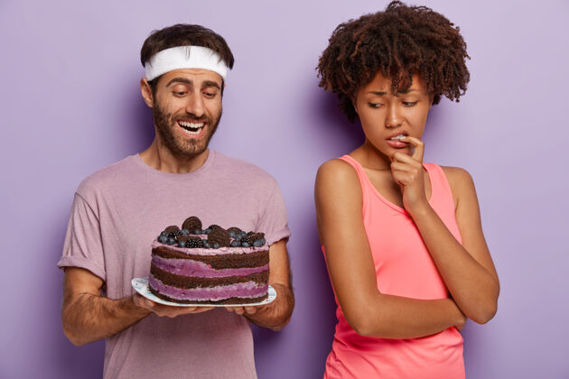 减肥男人给妻子提供美味的蛋糕迷惑不解的非裔美国女人回到丈夫身边 诱惑地看着甜点 避免垃圾食品 为了保持健康穿着运动服减肥 卡路里决定饮食拒绝
