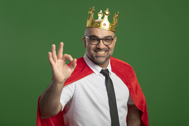 皇冠身穿红色斗篷 戴着皇冠眼镜的超级英雄商人面带微笑 站在绿白相间的墙壁上 露出ok标志眼镜人物穿着