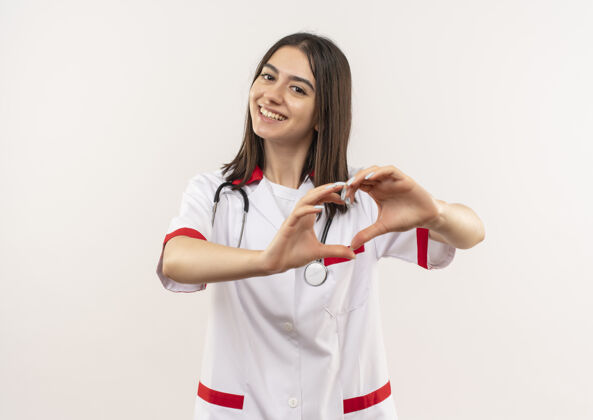 心脏年轻的女医生身穿白大褂 脖子上戴着听诊器 站在白墙上露出友好微笑的心形人外套脖子