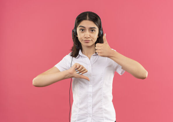 年轻人一个穿着白衬衫戴着耳机的年轻女孩 站在粉色的墙上 朝着前面竖起大拇指看公民女性