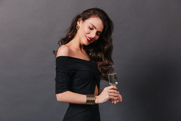红色唇膏优雅的女人 卷曲的长发 穿着时髦的黑色连衣裙 在深色背景下摆出香槟的姿势金手镯黑发闪耀
