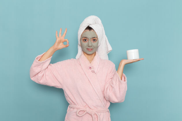 年轻人正面图：年轻女性 穿着粉色浴衣 脸上戴着口罩 蓝色桌上拿着面霜淋浴 清洁美容自我护理面霜人桌子自理