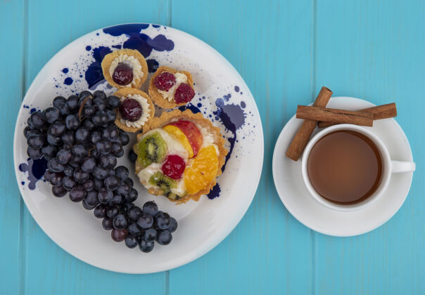 景观水果蛋糕和葡萄在盘子里的俯视图 以及一杯茶和肉桂茶碟在蓝色背景上的俯视图茶甜点肉桂
