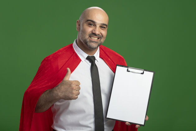 拇指穿红斗篷的超级英雄商人展示着剪贴板 空白的页面看着前面微笑着竖起大拇指站在绿色的墙上超级人脸