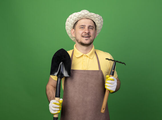人年轻的园丁穿着连体衣 戴着帽子 戴着工作手套 手里拿着小耙子和铲子 站在绿色的墙上 面带微笑地看着前方迷你连体衣人