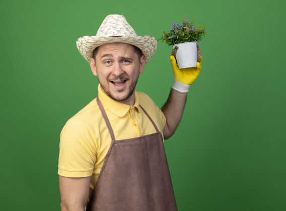 绿色年轻的园丁穿着连体衣 戴着帽子 戴着工作手套 手里拿着盆栽植物 站在绿色的墙上 面带微笑地看着前面看脸工作