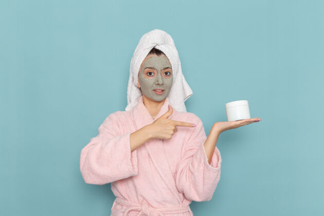 面部正面图：年轻女性 穿着粉色浴衣 脸上戴着口罩 拿着淡蓝色的面霜 淋浴 清洁 美容 自我护理年轻人成人面具