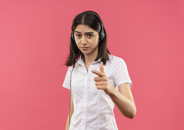 手指一个穿着白衬衫戴着耳机的年轻女孩 面带严肃的表情看着前面 手指指着前面站在粉红色的墙上女孩站着看