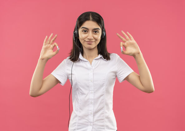 女孩穿着白衬衫 戴着耳机的年轻女孩 面带微笑 自信地站在粉红色的墙上 展示着ok标志年轻的手势微笑