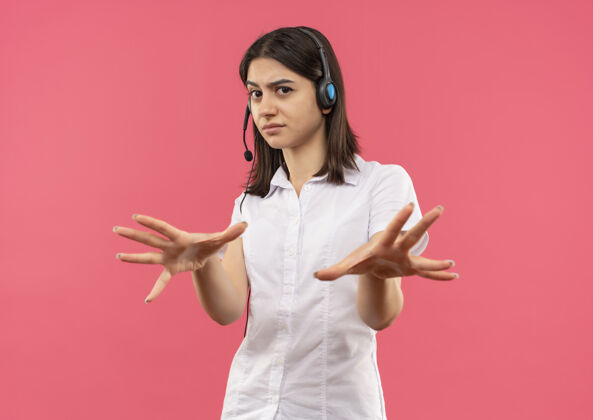 公民穿着白衬衫戴着耳机的年轻女孩站在粉色的墙上 用厌恶的表情做着防卫的手势手势站立麦克风