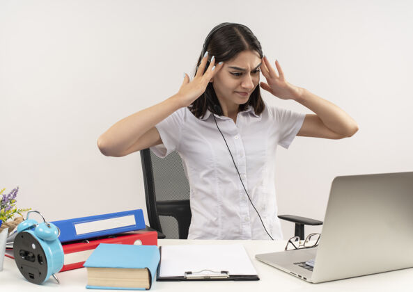 坐着一个穿着白衬衫戴着耳机的年轻女孩 两手抱着头坐在桌子旁 手里拿着文件夹和笔记本电脑 满脸困惑麦克风耳机衬衫
