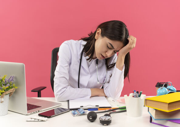 轮胎年轻的女医生穿着白大褂 脖子上戴着听诊器 看上去很累 她坐在桌边 粉色墙上挂着笔记本电脑 工作过度医疗保健过度工作周围