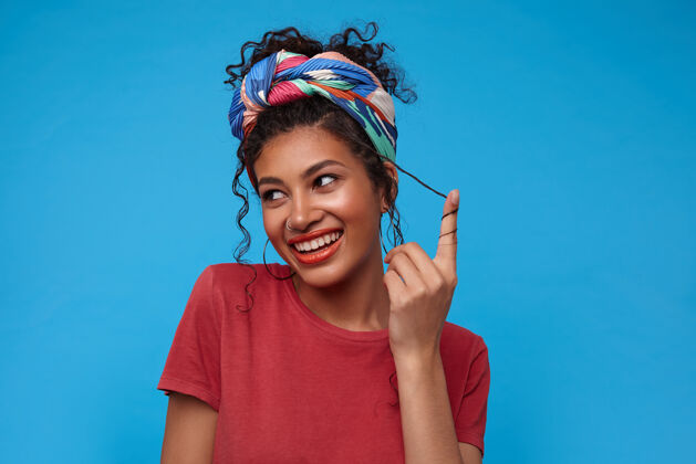 模特快乐的年轻迷人的黑发女子 戴着五颜六色的头巾 快乐地微笑着 把卷发扭在手指上 孤立在蓝色的墙上耳环黑发情感
