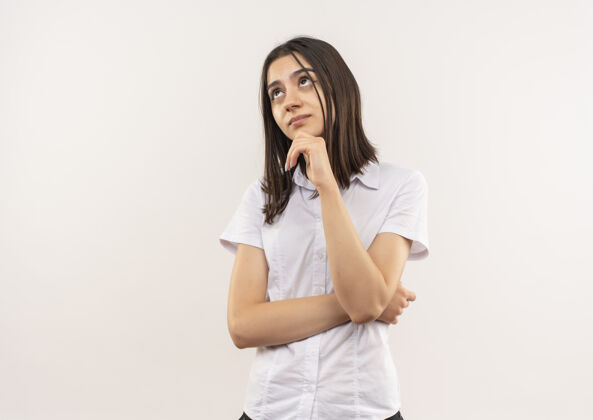手势穿着白衬衫的年轻女孩站在白墙上 手放在下巴上 困惑地看着一边靠边年轻人衬衫