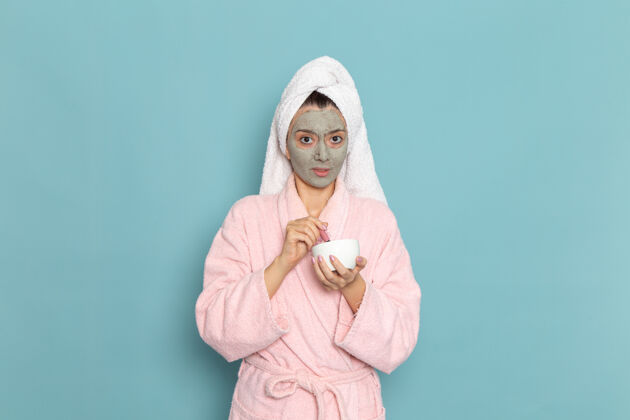 清洁正面图身着粉色浴袍的年轻女性淋浴后拿着面霜在蓝色墙壁上清洗美丽干净的水自我护理面霜淋浴人肖像前