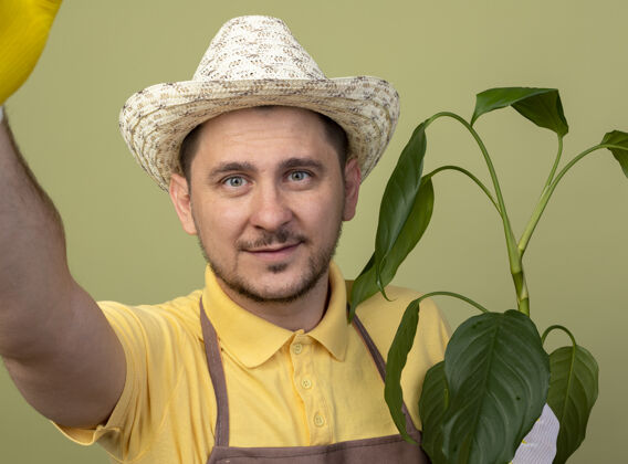 拿着年轻的园丁戴着工作手套 穿着连体衣 戴着帽子 拿着植物 面带微笑地看着前面 站在明亮的墙上植物微笑脸