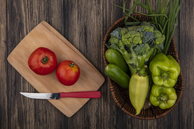 花椰菜俯视图：用刀在砧板上切西红柿 在木制背景上的篮子里放黄瓜 青椒 花椰菜和葱切餐午餐