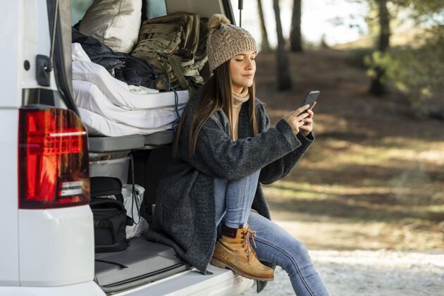 旅行坐在汽车后备箱里的一位女士的侧视图 她正在进行公路旅行并使用智能手机户外车辆乐趣