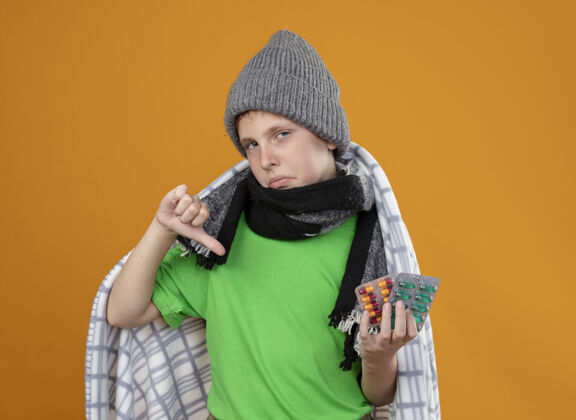 男孩生病的小男孩戴着暖和的帽子 围着围巾 裹着毯子 发烧 吃药 身体不适 站在橘色的墙上 拇指朝下拇指痛苦毯子