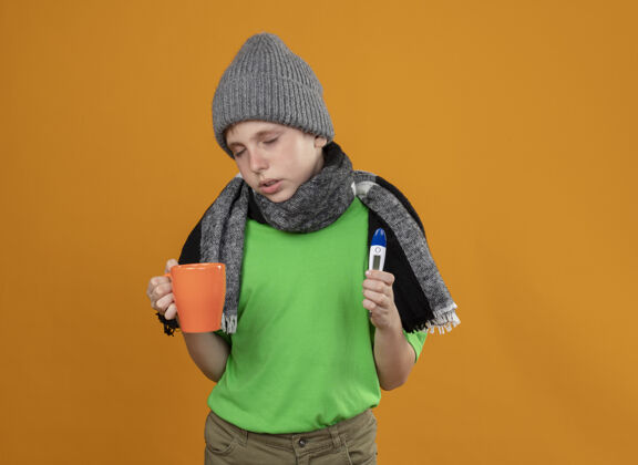 茶生病的小男孩穿着绿色t恤 戴着暖和的围巾 戴着帽子 手里拿着一杯热茶和温度计 站在橙色的墙上感到不舒服 不舒服和不开心不舒服温度计男孩