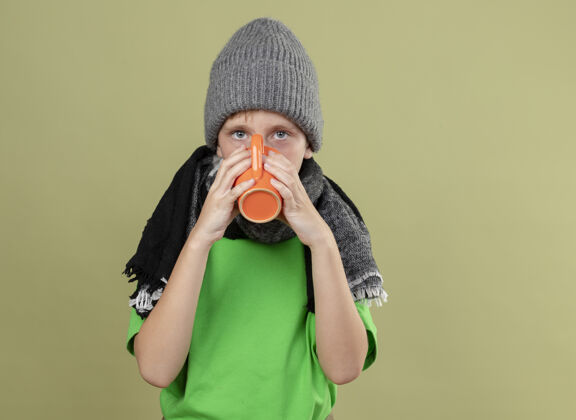 站生病的小男孩穿着绿色t恤 戴着暖和的围巾 戴着帽子 喝着热茶 忍受着寒冷站在轻墙上病男孩穿
