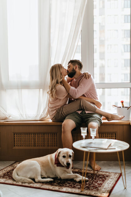 男性这对夫妇坐在窗台上亲吻穿着米色衣服的女孩拥抱着那个家伙 而他们的拉布拉多犬正躺在地毯上女朋友男朋友拉布拉多